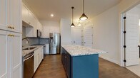 Located at 1081 N High St in Columbus's vibrant Short North neighborhood, this single-floor unit showcases a thoughtfully designed interior with contemporary finishes and a cohesive aesthetic. The open-concept kitchen serves as the centerpiece of the living space, featuring crisp white shaker cabinetry with gold hardware, a decorative arabesque tile backsplash, and granite countertops. A boldly colored navy blue island with matching granite provides generous prep and gathering space, anchored by industrial-style pendant lighting overhead. Stainless steel appliances, including a French door refrigerator, slide-in range, and built-in microwave, complete the kitchen's polished look. Rich, dark hardwood flooring flows throughout the main living and kitchen areas, lending warmth and continuity to the space. The bathroom is finished with large-format marble-look porcelain tile flooring, a glass-enclosed walk-in shower clad in classic subway tile with matte black hardware, a dark vanity with gold accents, and convenient access to a walk-in closet with built-in shelving. A carpeted bedroom features wainscoting-style wall paneling and a window that introduces natural light. The overall ambiance is modern yet inviting, with refined details and quality materials throughout.