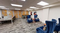 This single-floor space is a thoughtfully designed lounge or common area that blends bold aesthetics with functional comfort. The room is immediately eye-catching, anchored by a striking accent wall featuring a geometric orange-and-white pattern that adds energy and visual depth to the space. Complementing this are contemporary abstract art pieces displayed on adjacent walls, lending the area a creative and inspiring atmosphere.

The flooring consists of patterned carpet in neutral gray tones with subtle striping, providing a grounded contrast to the vibrant décor above. Seating is a standout feature, with plush royal blue wingback chairs accented by colorful striped throw pillows arranged around small orange side tables, creating intimate conversation areas throughout the room. Additional seating options include modern task chairs and a casual café-style table and chair grouping tucked near the accent wall, offering versatile use for both relaxed socializing and informal work.

Overhead, a suspended ceiling with recessed panel lighting ensures the space is evenly and brightly illuminated throughout. The overall design palette — combining deep blues, bold oranges, and crisp whites — creates a welcoming, dynamic environment well-suited for collaboration, relaxation, or informal gatherings.