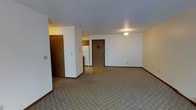 This single-floor space offers a straightforward and functional layout designed for comfortable everyday living. The main living area is generously sized, featuring neutral-toned carpet that extends throughout and white walls that create a bright, open feel. A textured ceiling adds a classic touch, while simple flush-mount lighting provides consistent illumination. The entryway is marked by a solid wood-toned door, complemented by matching wood-accented interior doors that lend a warm contrast to the light walls. Adjacent to the living area, the kitchen is visible through an open passage and includes wood-toned cabinetry and a white refrigerator, giving the space a clean, utilitarian character. The transition between the carpeted living area and the vinyl flooring of the kitchen and entryway zone is clearly defined, reinforcing the functional separation of spaces. A closet near the entry provides convenient storage access. Overall, the space presents a clean, move-in ready environment with a practical design, neutral color palette, and classic interior details suited to a wide range of occupants.
