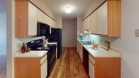 Located at 15300 37th Ave N in Plymouth, Minnesota, this single-floor unit offers a well-organized layout with distinct living, kitchen, sleeping, and bathroom areas. The galley-style kitchen is efficiently designed with ample upper and lower cabinetry finished in cream with warm wood trim, a double-basin sink, a dishwasher, and black appliances including a refrigerator, electric range, and over-the-range microwave. Durable wood-look flooring runs throughout the kitchen, providing a clean and easy-to-maintain surface. The living area is generously proportioned, featuring neutral wall-to-wall carpeting and a large sliding glass door dressed with vertical blinds that allows abundant natural light to fill the room. The transition between the carpeted living space and the wood-floored kitchen creates a natural separation of areas. The bedroom is a comfortable, carpeted room with a window offering an outdoor view and baseboard heating. The bathroom includes a wide vanity with an extended countertop, a large wall mirror, Hollywood-style lighting above, and warm-toned cabinetry below. Textured ceilings are present throughout the unit, and wood-toned baseboards add a consistent finishing detail across rooms. The overall layout is practical and well-suited for everyday living.