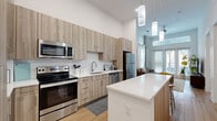 This thoughtfully designed one-bedroom, one-bathroom residence offers a contemporary open-concept layout across a single floor. The combined kitchen and living area serves as the heart of the home, featuring soaring ceilings that amplify the sense of space and allow natural light to pour in. The kitchen is outfitted with light wood cabinetry, a white quartz countertop island with seating for two, a subway tile backsplash, and a full suite of stainless steel appliances including a range, microwave, dishwasher, and refrigerator. Sleek pendant lighting adds warmth and visual interest overhead. The adjoining living area is bathed in natural light from full-length glass doors that open to a private balcony, with additional transom windows above enhancing the airy atmosphere. Light wood-tone flooring flows throughout, maintaining a clean, cohesive aesthetic. The bedroom is generously proportioned with high ceilings, neutral walls, and direct access to both the bathroom and a dedicated closet. The closet offers built-in shelving and hanging space for organized storage. The bathroom features a glass-enclosed walk-in shower with large-format white tile, a wood-toned vanity with a white countertop, and continued hardwood-style flooring, creating a spa-like retreat that complements the modern design sensibility found throughout the residence.
