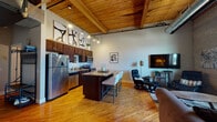 Located on Cesar E Chavez Ave in Grand Rapids, Michigan, this single-floor urban loft showcases a compelling blend of industrial character and modern comfort. The open-concept main living area immediately draws the eye upward to striking vaulted ceilings finished with warm wood planking and exposed structural beams, complemented by track lighting that adds an ambient glow throughout the space.

The kitchen is well-appointed with dark espresso cabinetry, stainless steel appliances including a full-size refrigerator, electric range, and microwave. A central island with a granite countertop and bar seating creates a natural gathering space that flows seamlessly into the living area. Hardwood floors extend throughout, reinforcing the cohesive, warm aesthetic.

The living room features a prominent exposed brick wall with large windows offering views of the Grand Rapids skyline, filling the space with natural light. A dedicated nook provides space for a desk and shelving, ideal for a home office setup.

The bedroom offers a quiet retreat with hardwood floors and connects conveniently to the bathroom, which features a granite-topped vanity with dark wood cabinetry and a walk-in shower. The overall ambiance balances industrial loft character with refined, livable details.