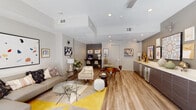 This thoughtfully designed single-floor communal lounge offers an open, flowing layout that seamlessly connects multiple functional zones. The main gathering area features a generously sized sectional sofa in warm taupe, anchored by a bold geometric yellow-and-grey area rug and a glass-top coffee table, creating an inviting space for relaxation. Natural light filters in through a large window, brightening the neutral grey and white palette throughout.

Along one wall, a sleek, elongated built-in credenza in dark woodgrain with a white quartz countertop serves as both a media console and bar area, complete with a stainless steel beverage cooler and an eclectic gallery wall of curated artwork. A wall-mounted flat-screen television is prominently positioned above the credenza for easy viewing from the lounge.

Adjacent to the lounge, a round dining table with mid-century modern chairs provides a casual yet refined dining area. Recessed lighting illuminates the space throughout, complemented by stylish pendant and table lamps.

Tucked into a dedicated alcove, a co-working station features a wall-mounted dark desk with two chairs, a desktop computer, and pendant cage lighting, ideal for productivity. Warm-toned wood-look flooring unifies the entire space, and carefully placed greenery and decorative accents add a welcoming, contemporary character throughout.