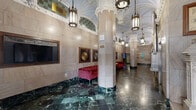 Located at 1509 Main Street in the heart of Dallas, Texas, the Kirby Building lobby is a stunning example of early 20th-century neoclassical architecture. The hallway makes an immediate impression with its soaring groin-vaulted ceiling, adorned with intricate plasterwork, Gothic-inspired detailing, and tiled accents in soft sage green — all illuminated by a series of elegant brass and glass lantern-style pendant fixtures that cast a warm, inviting glow throughout the space.

The floors are clad in richly veined green and black marble laid in a classic checkered pattern, lending the lobby a sense of timeless grandeur. Towering columns with ornately carved capitals line the corridor, reinforcing the building's distinguished architectural character. The walls feature smooth, warm-toned stone cladding trimmed in coordinating marble, maintaining a cohesive and refined aesthetic.

A seating area furnished with tufted red velvet sofas provides a striking contrast against the neutral palette, while framed botanical artwork adorns the walls. A prominent wall-mounted display panel, branded with the Kirby Building name, serves as a focal point. On the opposite end, polished brass elevator doors with Gothic detailing and arched entryways offer passage deeper into the building, completing this exceptional and historically significant lobby.