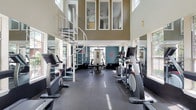 This fitness center is defined by its striking double-height ceilings and open, airy atmosphere. A prominent spiral staircase serves as an architectural centerpiece, rising to an upper mezzanine level that overlooks the main workout floor below, adding a dramatic sense of vertical scale to the space. Large windows line the walls throughout, flooding the interior with natural light and offering views of the surrounding outdoor environment, including glimpses of a pool area. Full-length mirrors along multiple walls further enhance the sense of openness and allow exercisers to monitor their form during workouts. The flooring throughout is durable rubberized gym matting, practical for high-traffic fitness use. The main floor is comprehensively equipped with a range of cardio machines, including treadmills, ellipticals, and stationary bikes, as well as a multi-function cable weight machine and a weight bench for strength training. A ceiling fan on the upper level provides additional air circulation. Glass double doors open directly to the outdoors, creating a seamless connection to the exterior. The teal accent walls add a vibrant pop of color, complementing the neutral tones of the overall design and contributing to an energizing workout environment.