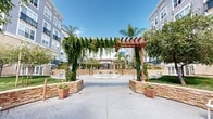 Located at 377 Santana Row in San Jose, California, this beautifully designed outdoor courtyard serves as a central gathering space within the Santana Row community. The space is defined by a striking wood pergola draped in cascading ivy, framing the main entry axis and drawing the eye toward a decorative central fountain that anchors the plaza. The paved areas feature intricate stamped and patterned stonework in circular and fan motifs, lending a refined, European-inspired aesthetic to the grounds. Stacked natural stone retaining walls curve gracefully through the space, delineating planting beds filled with ornamental shrubs, mature shade trees, and potted greenery that provide lush visual contrast against the hardscape. Tall, slender palm trees rise throughout the courtyard, reinforcing the warm California ambiance under open, sun-filled skies. Wrought-iron seating areas and decorative white benches are thoughtfully positioned to encourage relaxation and conversation. The courtyard is bordered by multi-story residential buildings with arched entryways, balconies, and large windows, including entrances labeled "Carneros" and "Alejandro." Black iron fencing with gated access adds a sense of security and definition to the perimeter, while low-profile landscape lighting enhances the space's functionality and atmosphere during evening hours.