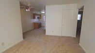 This single-floor residence offers a straightforward and functional layout designed for comfortable everyday living. The main living area is open and spacious, featuring light-toned carpeted flooring that extends throughout the space, contributing to a clean and neutral aesthetic. A ceiling fan with an integrated light fixture is centrally positioned overhead, providing both air circulation and ambient lighting.

The living area transitions seamlessly into the kitchen, which is outfitted with warm-toned wooden cabinetry arranged along the wall. The kitchen includes a sink set into a countertop with cabinetry above and below, offering ample storage and workspace. A window near the kitchen sink allows natural light to filter into the area, brightening the space. The kitchen also features a flush-mounted ceiling light for additional illumination.

The interior walls are finished in a soft, neutral cream tone, creating a bright and understated backdrop throughout. A door is visible along one wall of the living area, providing access to an additional room or closet. The overall ambiance of the space is simple, clean, and move-in ready, making it a practical and versatile option for a variety of living arrangements.