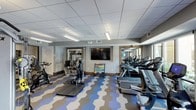 This fitness center at 4800 Marriott Dr in Raleigh, NC, offers a thoughtfully designed workout space that combines functionality with a modern aesthetic. The room is defined by its eye-catching geometric-patterned flooring in shades of blue, grey, and white, adding a dynamic visual element to the space.

The facility is well-lit with recessed panel lighting and suspended fluorescent fixtures throughout the drop-tile ceiling, ensuring bright, consistent illumination across the entire floor. A large flat-screen television is mounted on the central wall, providing entertainment during workouts.

The space is comprehensively equipped with a row of treadmills and elliptical machines along one wall, ideal for cardiovascular training. A dedicated cycling station features stationary bikes, while a step platform provides options for aerobic exercise. A color-coded dumbbell rack offers a range of free weights for strength training. Additional strength equipment is visible through a mirrored adjoining area, expanding the available workout options.

Large mirrors are strategically placed along the walls, enhancing the sense of openness and allowing users to monitor their form. The neutral grey walls and clean architectural trim contribute to a polished, welcoming atmosphere throughout the fitness center.