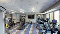 This fitness center at 4800 Marriott Dr in Raleigh, NC, offers a thoughtfully designed workout space that combines functionality with a modern aesthetic. The room is defined by its eye-catching geometric-patterned flooring in shades of blue, grey, and white, adding a dynamic visual element to the space.

The facility is well-lit with recessed panel lighting and suspended fluorescent fixtures throughout the drop-tile ceiling, ensuring bright, consistent illumination across the entire floor. A large flat-screen television is mounted on the central wall, providing entertainment during workouts.

The space is comprehensively equipped with a row of treadmills and elliptical machines along one wall, ideal for cardiovascular training. A dedicated cycling station features stationary bikes, while a step platform provides options for aerobic exercise. A color-coded dumbbell rack offers a range of free weights for strength training. Additional strength equipment is visible through a mirrored adjoining area, expanding the available workout options.

Large mirrors are strategically placed along the walls, enhancing the sense of openness and allowing users to monitor their form. The neutral grey walls and clean architectural trim contribute to a polished, welcoming atmosphere throughout the fitness center.