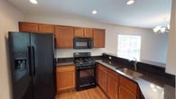 This single-floor home offers a thoughtfully designed layout with a variety of functional and comfortable living spaces. The kitchen is a standout feature, outfitted with warm medium-tone wood cabinetry, dark granite-style countertops, and a full suite of black appliances including a gas range, over-the-range microwave, and a side-by-side refrigerator with an ice and water dispenser. Recessed lighting and a window above the sink provide ample illumination, while wood-look flooring adds warmth underfoot. A peninsula counter seamlessly connects the kitchen to the adjacent living and dining area, which is bathed in natural light from large windows dressed with blinds. An elegant chandelier anchors the dining space, and the same wood-look flooring continues throughout, creating a cohesive flow. The bedrooms are carpeted for comfort, featuring neutral walls, generous windows that invite natural light, and well-sized closets — one bedroom includes a spacious walk-in closet with built-in shelving and hanging rods. The bathroom is clean and practical, offering a combined tub and shower, a wood-vanity with a dark countertop and undermount sink, a large mirror, and wood-look flooring. Throughout the home, a neutral color palette and consistent finishes create a bright, welcoming ambiance.