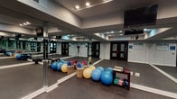 This aerobics room is a well-appointed fitness space designed to accommodate a wide range of exercise activities. The room is defined by its floor-to-ceiling mirrored walls that line multiple sides, creating a sense of expansive openness while allowing participants to monitor their form and technique during workouts. The mirrors also reflect the recessed ceiling lighting, keeping the space bright and well-illuminated throughout.

The flooring consists of durable, dark rubber matting, which provides both cushioning and traction ideal for aerobic and fitness activities. A raised ceiling with a stepped architectural detail adds visual interest and contributes to the room's airy feel. A wall-mounted flat-screen television is positioned for clear visibility across the room, supporting instructed classes or individual workout sessions.

The space is equipped with an assortment of fitness tools, including stability balls in multiple sizes and colors, foam rollers, hand weights, and a small equipment rack. Entry is provided through a set of double doors with glass panel inserts, lending a polished, professional appearance to the entrance. The neutral gray walls and clean white baseboards maintain a modern, clean aesthetic throughout. Overall, this aerobics room offers a functional, versatile environment suited for group fitness classes, personal training, and a broad range of exercise disciplines.