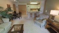 This single-floor residence offers a thoughtfully designed open-concept layout that seamlessly connects the living, dining, and kitchen areas into one cohesive and functional space. The living area is finished with plush wall-to-wall carpeting, creating a warm and comfortable atmosphere, and is furnished with a patterned accent chair and a glass-top coffee table. Adjacent to the living space, a defined dining area accommodates a dark wood table and chair set, ideal for everyday meals or casual gatherings. A sliding closet with natural wood-toned doors provides convenient storage nearby. The kitchen is positioned at the heart of the home, featuring white cabinetry, a full-size refrigerator, and a raised breakfast bar with two wooden barstools — perfect for quick meals or casual conversation. Recessed lighting and a built-in overhead fixture illuminate the kitchen efficiently, while track lighting accents the living and dining zones. The clean, neutral color palette throughout — from the white cabinetry to the light carpeting — lends the space a bright and welcoming feel. Overall, this residence combines practical storage solutions with an open, livable floor plan suited to a variety of lifestyles.