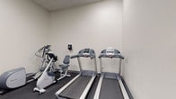 This fitness room is a purpose-built exercise space designed to support a well-rounded workout routine. The room features durable rubber flooring throughout, providing a safe and practical surface for all types of physical activity. The walls are finished in a neutral off-white tone, contributing to a clean and functional aesthetic, while a drop tile ceiling with recessed lighting ensures the space is evenly and adequately lit.

The cardio section is well-appointed, housing two treadmills and an elliptical trainer, offering users multiple options for aerobic exercise. A stationary recumbent-style bike rounds out the cardio offerings, catering to a variety of fitness preferences and needs. A wall-mounted hand sanitizer dispenser is conveniently located nearby, promoting hygiene throughout workouts.

Along one wall, a multi-tier storage rack holds an assortment of fitness accessories including medicine balls of varying weights, push-up handles, a foam roller, a yoga mat, and a stability ball — making the room suitable for strength training and flexibility exercises as well. A small pass-through window connects to an adjacent area, adding a subtle architectural detail to the otherwise straightforward layout. The overall design of this fitness room prioritizes practicality and utility, offering a compact yet comprehensive environment for daily exercise.