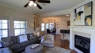 This two-story home offers a thoughtfully designed interior that balances comfort and style across its multiple levels. The main living area showcases rich hardwood flooring and classic crown molding throughout, lending a polished, timeless character to the space.

The living room is anchored by a ceiling fan, a cozy fireplace with a crisp white mantel, and plush seating arranged around a rounded ottoman coffee table. Natural light filters in through multiple windows, enhancing the warm, inviting atmosphere. A patterned accent chair and layered area rugs add texture and depth to the room.

The living space flows seamlessly into a formal dining area, defined by an elegant chandelier suspended above a dark wood dining table and upholstered chairs. Decorative plate arrangements on the wall add a curated, artistic touch to the dining space.

Adjacent to the dining area, a glimpse of the kitchen reveals dark cabinetry, bar-height seating, and additional natural light — suggesting a well-equipped culinary space with an open connection to the entertaining areas. The cohesive neutral color palette of warm whites and grays ties the entire main floor together, creating an atmosphere that is both functional and aesthetically refined.