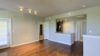 This single-story space features an open-concept design that seamlessly connects the living area to the kitchen. Rich, dark hardwood floors run throughout the main living space, providing a warm contrast to the soft, neutral gray-green walls and bright white trim and baseboards. The living area is illuminated by a track lighting system mounted on the ceiling, ensuring well-distributed light across the room. A peninsula-style kitchen pass-through with a granite or stone countertop visually separates the kitchen from the living area while maintaining an open, airy feel. The kitchen itself, visible through the pass-through, features stainless steel appliances, white cabinetry, and matching track lighting. HVAC vents are integrated cleanly into the ceiling for climate control. A doorway from the living area leads to additional rooms, including what appears to be a bedroom with carpeted flooring visible in the hallway. White panel doors are used consistently throughout, adding to the clean, cohesive aesthetic. The overall ambiance is modern and move-in ready, with a neutral palette that lends itself well to a variety of interior design styles.