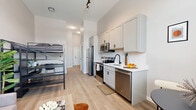 Located at 110 Cornell St in Kingston, New York, this thoughtfully designed studio unit makes smart use of its single-floor layout. The open-plan kitchen and living area serves as the heart of the space, featuring light gray shaker-style cabinetry, a white countertop, stainless steel appliances including a refrigerator, dishwasher, range, and microwave, and a black matte faucet over a clean undermount sink. Light wood-tone flooring flows throughout, enhancing the bright, airy feel. The living area accommodates a sectional sofa and coffee table, with high ceilings that amplify the sense of space — a notable standout being the loft sleeping platform accessed via a built-in staircase with integrated shelving and a dedicated desk below, creating an efficient live-work zone. The bathroom is clean and contemporary, outfitted with a gray shaker-style vanity, white countertop, black hardware, a large framed mirror with lighted accents, and wood-look tile flooring. Neutral tones and consistent finishes throughout give the unit a cohesive, modern aesthetic. Situated in Kingston's vibrant community, this unit is an ideal option for those seeking efficient, well-appointed urban living in the Hudson Valley.