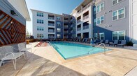 Located at 2200 Silver Crescent Dr in Charlotte, NC, this outdoor amenity space is designed to deliver a resort-style experience within a modern residential community. The centerpiece is a generously sized swimming pool with crystal-clear water, framed by an expansive travertine tile sundeck that provides ample space for lounging. Rows of poolside chaise lounges offer residents a comfortable place to relax and enjoy the open sky, while a decorative wooden geometric wall art installation adds an artistic focal point to the poolside atmosphere.

The surrounding multi-story residential building features a contemporary design with a striking combination of blue and light gray exterior siding, large windows, and private balconies that overlook the pool area. Double glass-panel entry doors lead into the amenity building, allowing natural light to flow through and maintaining a visual connection to the pool.

The space is thoughtfully secured with a gated black iron perimeter fence, complete with clearly marked exit points for safety. Lush landscaping with tropical-style plants lines the pathways, enhancing the welcoming ambiance. A dedicated pool equipment room is also present on-site, ensuring the facility is well-maintained. The overall atmosphere is bright, open, and contemporary, ideal for outdoor recreation and relaxation.