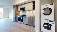 This thoughtfully designed 1-bedroom, 1-bathroom unit presents a clean, modern aesthetic throughout its single-floor layout. Light wood-tone laminate flooring flows seamlessly across all living areas, creating a cohesive and airy feel. The open-concept living and kitchen area is anchored by a floor-to-ceiling sliding glass door that floods the space with natural light and provides outdoor views of surrounding greenery.

The kitchen is efficiently designed with two-tone cabinetry — warm wood-grain lower cabinets paired with light cream uppers — complemented by a white subway tile backsplash and under-cabinet lighting. A full suite of stainless steel appliances includes an electric range, over-the-range microwave, dishwasher, and refrigerator. A stacked washer and dryer unit is conveniently located adjacent to the kitchen.

The bedroom is a well-proportioned retreat featuring light-filled windows with outdoor views, a built-in sliding door closet, and the same warm flooring found throughout. The bathroom offers a clean, contemporary finish with large-format tile surrounding a soaking tub with shower, a wood-grain vanity, and a mirrored medicine cabinet. White walls and crisp trim complete the bright, modern character of this well-appointed space.