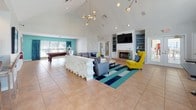 Located at 650 W Flintlake Ct in Myrtle Beach, SC, this single-floor clubhouse is a well-appointed community gathering space designed with a bold, contemporary aesthetic. The main lounge is the heart of the facility, boasting soaring vaulted ceilings that create an airy, open atmosphere. A striking sputnik chandelier serves as a focal point overhead, while large floor-to-ceiling windows with teal drape accents flood the space with natural light and provide views of the surrounding community. The lounge features a generous sectional sofa and accent seating in vibrant yellow, anchored by a bold striped area rug in teal, navy, and neutral tones over warm tile flooring. A tiled fireplace with built-in shelving and a mounted flat-screen TV creates an inviting entertainment wall. Adjacent to the lounge, a dedicated billiards area offers recreational space for residents. The kitchen and bar area is a standout feature, centered around a curved, navy-paneled bar with a marble-top counter and modern chrome barstools, all set against teal accent walls and contemporary artwork. The full kitchen behind the bar includes stainless steel appliances. A separate lounge nook with bistro-style tables and seating near the windows provides a quieter setting for conversation or casual work.