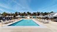 Located in Daphne, Alabama, this single-story residential community along Alabama 181 offers a well-appointed outdoor living experience in a serene, wooded setting. The centerpiece of the community is a generously sized resort-style swimming pool with crystal-clear blue water, surrounded by a spacious concrete deck designed for relaxation and recreation. The pool area is outfitted with multiple lounge chairs and chaise seating arrangements, providing ample space for sunbathing and leisure. Vibrant teal umbrella-shaded cabanas are positioned throughout the deck, offering comfortable shaded retreats on sunny days. A wrought iron perimeter fence encloses the pool area, ensuring both safety and an open, airy aesthetic. In the background, the community's residential buildings are visible — featuring classic pitched rooflines and a cohesive architectural style that blends harmoniously with the natural surroundings. Tall, mature trees form a lush green backdrop, lending the space a private, park-like atmosphere. The overall ambiance is bright and welcoming, with the wide-open Alabama sky completing the sense of space and tranquility that defines this community's outdoor amenity area.