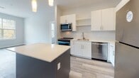 Located at 1 Royal Dr in Somersworth, New Hampshire, this single-floor unit offers a thoughtfully designed layout with modern finishes and a bright, clean aesthetic throughout. The open living area features light gray walls and smooth hardwood-style flooring, with a door and windows providing natural light and access to an outdoor balcony area surrounded by lush greenery.

The contemporary kitchen is a standout feature, equipped with crisp white cabinetry — both upper and lower — paired with light quartz countertops and stainless steel appliances, including a refrigerator, range, microwave, and dishwasher. A kitchen island with a dark base adds contrast and extra workspace, while pendant lighting overhead completes the modern look. Light wood-style flooring ties the space together seamlessly.

The bedrooms are carpeted in neutral tones and bathed in natural light from large windows that frame views of the surrounding trees and outdoor balcony. The soft color palette of light blue-gray walls creates a calm and restful atmosphere in each room.

A well-appointed hallway connects the living areas to the bedrooms and bathroom, which features a clean white tub-shower combination with a chrome towel bar and light tile flooring. The overall design of this unit emphasizes functionality and modern simplicity, making it a comfortable and well-organized living space.