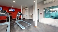 Located at 44 Salix Ct in Baltimore, Maryland, this fitness center is a single-floor facility designed to support a well-rounded workout experience. The space is divided into distinct workout zones, creating an organized and functional layout for users.

The cardio area is a standout section, featuring multiple treadmills and stationary bikes arranged on durable, speckled rubber flooring. A bold red accent wall serves as a visual focal point, with wall-mounted flat-screen televisions positioned for easy viewing during workouts. Recessed lighting keeps the area bright and energizing.

The strength and functional training area offers a multi-station cable machine and an assortment of stability balls in various sizes, neatly stored on a dedicated rack. This section benefits from an abundance of natural light streaming through large windows and a glass entry door, which also provides views of the surrounding greenery outdoors.

The facility includes practical amenities such as a dual-level water fountain and bottle-filling station, conveniently located near a posted Fitness Guidelines sign. A wall-mounted mini-split HVAC unit ensures year-round climate comfort. Structural columns are cleanly integrated into the layout, and the overall design uses a neutral white and gray palette, accented by teal and red details, giving the space a modern, clean aesthetic.