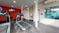 Located at 44 Salix Ct in Baltimore, Maryland, this fitness center is a single-floor facility designed to support a well-rounded workout experience. The space is divided into distinct workout zones, creating an organized and functional layout for users.

The cardio area is a standout section, featuring multiple treadmills and stationary bikes arranged on durable, speckled rubber flooring. A bold red accent wall serves as a visual focal point, with wall-mounted flat-screen televisions positioned for easy viewing during workouts. Recessed lighting keeps the area bright and energizing.

The strength and functional training area offers a multi-station cable machine and an assortment of stability balls in various sizes, neatly stored on a dedicated rack. This section benefits from an abundance of natural light streaming through large windows and a glass entry door, which also provides views of the surrounding greenery outdoors.

The facility includes practical amenities such as a dual-level water fountain and bottle-filling station, conveniently located near a posted Fitness Guidelines sign. A wall-mounted mini-split HVAC unit ensures year-round climate comfort. Structural columns are cleanly integrated into the layout, and the overall design uses a neutral white and gray palette, accented by teal and red details, giving the space a modern, clean aesthetic.