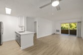 This thoughtfully designed one-bedroom, one-bathroom unit offers a compact yet efficient single-level layout. The open-concept kitchen and living area forms the heart of the space, finished with light wood-style flooring that runs throughout and crisp white walls that enhance the overall brightness. The kitchen features a central island with a white quartz countertop and an integrated sink with a modern pull-down faucet, complemented by white cabinetry, a skylight overhead, and a full suite of stainless steel appliances. The living area is generously proportioned for its scale, equipped with a ceiling fan and a wall-mounted heating and cooling unit. Sliding glass doors open to a balcony, allowing natural light to fill the room and offering views of surrounding greenery. The bedroom is a comfortable, well-lit room with a large window dressed in vertical blinds and baseboard heating. The bathroom maintains the clean aesthetic of the unit, featuring a white vanity with ample counter space, wood-style flooring, and neutral walls. Textured ceilings are consistent throughout, adding subtle character to this clean, move-in-ready space.