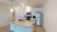 This single-story residence is thoughtfully designed with a cohesive, contemporary aesthetic throughout. The kitchen serves as a central highlight, featuring a large granite-topped island with an integrated sink, crisp white cabinetry with brushed nickel hardware, a light blue subway tile backsplash, and a full suite of stainless steel appliances including a refrigerator, electric range, and over-the-range microwave. Wood-look flooring runs through the kitchen, complemented by pendant lighting above the island that enhances the bright, functional workspace.

The living area transitions seamlessly from the kitchen and is finished with plush neutral carpeting and a ceiling fan with light fixture, creating a comfortable, well-ventilated environment. The bedroom continues this approach, offering generous proportions with soft carpeting, a walk-in closet equipped with wire shelving, and direct access to the adjoining bathroom.

The bathroom is well-appointed, featuring a dual-sink vanity with granite countertops, white cabinetry, a large mirror with overhead lighting, and wood-look tile flooring. A dedicated in-unit laundry closet houses a full-size washer and dryer with overhead wire shelving for additional storage. Neutral tones and consistent finishes throughout contribute to a clean, cohesive living environment.