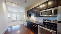 This single-floor unit is designed with a cohesive, contemporary aesthetic throughout. The kitchen serves as a standout feature, showcasing rich dark espresso cabinetry that extends to the ceiling, offering generous storage capacity. A mosaic tile backsplash in neutral tones complements the dark lower cabinets and dark countertops, while a full suite of stainless steel appliances—including a refrigerator, electric range, built-in microwave, and dishwasher—adds a polished, functional finish. Hardwood flooring runs throughout the kitchen and flows into the adjacent living area, which is illuminated by a ceiling fan and benefits from natural light provided by multiple windows fitted with horizontal blinds. The living space transitions seamlessly from the kitchen, creating an open, connected layout. The bedroom is carpeted for comfort and features multiple windows that allow natural light to fill the room, along with a ceiling fan. The bathroom includes a soaking tub with tile surround, tile flooring, and direct access to a walk-in closet with built-in shelving, providing practical and well-organized storage. Track lighting in the kitchen area enhances visibility and adds a modern touch to the overall design.