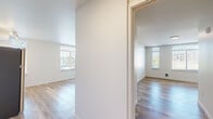 Located at 3300 NE 125th St in Seattle's Meadowbrook neighborhood, this one-bedroom, one-bathroom unit offers a thoughtfully designed single-floor layout. Light hardwood-style flooring runs throughout, complementing the bright, neutral palette of crisp white walls and clean-lined finishes.

The open kitchen and living area form the heart of the unit. The kitchen features white upper cabinetry, two-tone lower cabinets, a stainless steel refrigerator, microwave, and an in-unit washer, all positioned beneath track lighting. The living area receives ample natural light through large windows and includes a private entry door with a sidelight panel and transom window that opens to a small balcony.

The bedroom is generously sized, offering two large windows with views of the surrounding Seattle greenery and neighborhood, providing a bright and airy atmosphere. The room offers solid proportions well-suited for full bedroom furnishings.

The bathroom is clean and functional, featuring a white vanity with a modern basin sink, a large mirror, and wood-look flooring that ties in with the rest of the unit.

Throughout the space, abundant natural light, a consistent modern aesthetic, and efficient use of layout make this a practical and comfortable urban residence in the north Seattle area.