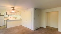This single-floor residential space offers a straightforward, functional layout designed for comfortable everyday living. The main living area is finished with wall-to-wall carpeting that adds warmth underfoot, while the white-painted walls create a bright, neutral backdrop throughout. A ceiling fan is centrally positioned in the living area, providing both air circulation and ambient lighting.

A partial dividing wall separates the living space from an adjacent area, adding a sense of structure to the open floor plan without fully closing off the rooms. A pass-through counter connects the kitchen to the living area, allowing for easy interaction between the two spaces. The kitchen features overhead cabinetry with a dark-framed, grid-style design that provides a subtle visual contrast against the light surroundings.

Closet storage is accessible directly from the main living area via a white panel door, offering practical organization space. A floor-level ventilation register is visible along the baseboard, indicating a central HVAC system for climate control. A doorway at the far end of the space leads to additional rooms, suggesting further functional areas beyond what is immediately visible. Overall, the space presents a clean, utilitarian design with easy-to-maintain finishes and a cohesive, light-toned aesthetic.