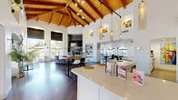 Located at 14435 South 48th Street in Phoenix, AZ, this single-floor community space makes an immediate impression with its dramatic vaulted ceilings adorned with exposed wood beams and warm tongue-and-groove paneling. The open-plan layout flows seamlessly across rich dark hardwood flooring, connecting multiple functional zones into one cohesive, inviting environment.

The kitchen is a standout feature, outfitted with white shaker cabinetry, a marble-style quartz countertop island with bar seating, stainless steel appliances including a professional range with hood, a French door refrigerator, and a marble tile backsplash. Elegant pendant lighting hangs above the island, and sliding glass doors provide direct access to an outdoor patio.

The main gathering area centers around high communal tables and modern bar-height chairs, with a stone-surround fireplace and wall-mounted television serving as a focal point. Arched windows and oversized roller shades frame views of the lush Phoenix landscape, flooding the space with natural light. A cozy lounge nook features a curved sectional sofa accented with colorful throw pillows, surrounded by tall windows and greenery.

A separate billiards area with a full-size pool table and a dedicated dining zone with arched window views round out the layout, making this a well-appointed, multi-use space designed for both community engagement and relaxed recreation.