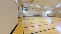 Located at 5617 111 St E in Puyallup, Washington, this single-floor indoor sports facility is designed to accommodate a range of athletic and recreational activities. The centerpiece of the space is a full-size basketball court finished with polished hardwood flooring, clearly marked with regulation court lines in black and red. Soaring ceilings rise dramatically overhead, creating an expansive, open atmosphere that is well-suited for active use. Natural light streams in through a series of skylights positioned at the ceiling's peak, supplemented by recessed and pendant lighting fixtures that ensure consistent illumination throughout the court. A basketball hoop is mounted on one end of the court, underscoring the facility's athletic purpose. Along one wall, a raised platform with built-in upholstered seating provides a dedicated spectator or staging area, flanked by built-in storage cabinetry with panel doors. Large windows on one side of the court allow additional natural light to filter in while offering views of the surrounding greenery. A carpeted staircase with dual handrails provides access between levels, finished with warm-toned walls and an emergency exit sign above the landing. The overall aesthetic is clean, functional, and well-maintained, making this facility a versatile space for sports, events, or community programming.