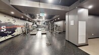 Located at 2475 Hurontario Street in Mississauga, Ontario, this fitness centre offers a well-appointed exercise space designed to support a full range of workout routines. The layout is thoughtfully organized into distinct zones, accommodating both cardio and strength training needs.

The cardio area is generously equipped with multiple treadmills and elliptical cross-trainers, positioned alongside a full-length mirrored wall that amplifies the sense of space and allows users to monitor their form. Durable speckled rubber flooring runs throughout this section, providing excellent grip and impact absorption.

The strength training area features a multi-function cable machine, a set of dumbbells on a tiered rack, an adjustable weight bench, and a stability ball, offering a comprehensive free weights zone. A wall-mounted punching pad adds an option for functional and martial arts-style training.

A bold, graphic feature wall with a dynamic fitness-themed mural serves as a striking visual focal point, adding energy to the environment. Track lighting and recessed ceiling fixtures provide bright, even illumination throughout the space. The dark ceiling and grey walls create a sleek, modern aesthetic that is both motivating and functional.

Overall, this fitness centre is a well-rounded facility suited for residents or building occupants seeking convenient, on-site access to professional-grade exercise equipment.