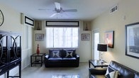 This single-floor space presents a thoughtfully designed living area that balances comfort and style. The living room serves as the centerpiece, featuring a sleek black leather sofa accented with contrasting geometric throw pillows, creating a bold yet inviting atmosphere. A ceiling fan with a classic multi-blade design is centrally mounted, promoting air circulation throughout the room. Large windows dressed with vertical blinds and a structured valance allow generous natural light to fill the space, enhancing its open and airy feel. The neutral, light-toned walls and carpeted flooring provide a clean backdrop that complements the darker furnishings. Decorative elements are carefully arranged throughout, including framed wall art, sculptural accent pieces, and matching side tables that lend the space a curated, intentional aesthetic. A stylish floor lamp with a bold black shade adds a layer of ambient lighting, while a glass-topped coffee table contributes a touch of modern elegance. An entry door with a sidelight window is visible, connecting the living area to the outside and allowing additional natural light to enter. Overall, the space exudes a modern, monochromatic character with a warm and livable ambiance.