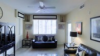 This single-floor space presents a thoughtfully designed living area that balances comfort and style. The living room serves as the centerpiece, featuring a sleek black leather sofa accented with contrasting geometric throw pillows, creating a bold yet inviting atmosphere. A ceiling fan with a classic multi-blade design is centrally mounted, promoting air circulation throughout the room. Large windows dressed with vertical blinds and a structured valance allow generous natural light to fill the space, enhancing its open and airy feel. The neutral, light-toned walls and carpeted flooring provide a clean backdrop that complements the darker furnishings. Decorative elements are carefully arranged throughout, including framed wall art, sculptural accent pieces, and matching side tables that lend the space a curated, intentional aesthetic. A stylish floor lamp with a bold black shade adds a layer of ambient lighting, while a glass-topped coffee table contributes a touch of modern elegance. An entry door with a sidelight window is visible, connecting the living area to the outside and allowing additional natural light to enter. Overall, the space exudes a modern, monochromatic character with a warm and livable ambiance.