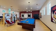 Located at 2201 Greenville Blvd in Greenville, NC, this single-floor amenity space combines recreation, relaxation, and productivity in one well-appointed environment. The open layout is anchored by a full-size pool table with a bright blue felt surface, positioned beneath a striking multi-arm sputnik chandelier that serves as a focal point of the room. Light hardwood flooring runs throughout, complementing the clean, modern aesthetic.

The lounge area features a bold deep crimson accent wall adorned with a large-scale geometric black-and-white art installation framing dual mounted flat-screen televisions. Comfortable gray sectional sofas and vibrant red accent chairs are arranged to encourage social gathering, while a marble-topped coffee table adds a refined touch.

A dedicated co-working zone offers individual workstations with wood-topped desks, illuminated by warm yellow pendant lighting against a backdrop of soaring ceilings and tall windows that draw in abundant natural light. The high ceilings throughout the space enhance its open, airy feel.

Bold pop-art wall pieces and curated decor add personality and energy to the space. Teal-painted doors provide additional architectural interest, while open shelving near the lounge area offers functional display space. The overall ambiance is lively, modern, and thoughtfully designed to serve as a versatile community gathering hub.