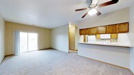 This single-floor residence offers a practical and comfortable open-concept layout designed for easy everyday living. The spacious main living area is finished with plush wall-to-wall carpeting and neutral-toned walls, creating a warm and inviting atmosphere. A ceiling fan with integrated lighting serves as a central fixture, providing both air circulation and ambient light throughout the space.

The kitchen is positioned along one side of the open floor plan and features rich, warm-toned wood cabinetry with raised-panel doors offering generous storage. White appliances, including a refrigerator and built-in microwave, complement the clean aesthetic. A raised breakfast bar counter separates the kitchen from the living area, making it an ideal spot for casual dining or conversation while cooking.

Natural light filters into the living area through a sliding glass door, which also provides access to the outdoors and contributes to the airy, open feel of the space. A hallway visible from the main area connects to additional rooms, suggesting a thoughtful layout that balances open communal space with private areas. Overall, the design is functional and welcoming, suited for comfortable everyday living.