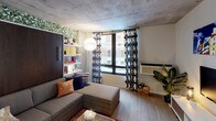 This single-floor studio is a masterclass in efficient, stylish living. The main living area is anchored by a sleek murphy bed with a rich dark wood finish, seamlessly integrated into a wall unit with built-in open shelving, allowing the space to function as both a comfortable lounge and sleeping quarters. A bold botanical wallpaper accent wall lends the area a lively, urban character, while the exposed concrete ceiling adds an industrial edge. Large windows dressed with geometric-patterned curtains invite natural light and offer views of the surrounding cityscape. The kitchen is compact yet fully functional, featuring stainless steel appliances including a full-size refrigerator and over-the-range microwave, wood-grain lower cabinetry, a stone countertop, and open upper shelving for added storage. The bathroom makes a strong design statement with bold blue and white horizontal stripes adorning the walls, complemented by a combined tub and shower with a rainfall showerhead. Wood-look flooring flows throughout the unit, tying the spaces together cohesively. A stacked in-unit washer and dryer rounds out the practical amenities of this well-appointed studio.