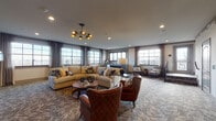 This thoughtfully designed single-floor communal lounge space offers a warm and sophisticated atmosphere, blending contemporary style with inviting comfort. The open layout is anchored by a generously sized carpeted living area featuring a large sectional sofa, richly upholstered cognac leather armchairs, and a round wooden coffee table — all centered beneath a striking multi-arm chandelier with warm brass accents.

A focal wall showcases a modern linear fireplace framed by floor-to-ceiling wood-paneled surround and a mounted flat-screen television above, creating an ideal gathering spot. Adjacent to the fireplace, nesting metal side tables and additional seating provide a flexible arrangement for various group sizes.

Large multi-pane windows line multiple walls, flooding the space with natural light and offering expansive outdoor views. Layered textured carpeting and neutral-toned walls contribute to the space's refined, contemporary aesthetic. Sheer and structured drapery panels add a polished finishing touch throughout.

A dedicated wet bar area features a deep-set sink, stainless steel cabinetry, and wood-look wall paneling, ideal for entertaining. The space also includes a transitional entry zone with stair access, additional lounge seating, restroom facilities, and recessed lighting throughout, completing this well-appointed communal retreat.