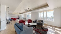 Located atop a Philadelphia building in the 19123 zip code, this common area roof lounge offers a sophisticated and well-appointed gathering space designed for comfort and socializing. The lounge is organized into distinct zones across an elongated open floor plan, all unified by warm herringbone-patterned hardwood flooring that adds a refined architectural detail throughout.

The main seating area is furnished with bold red leather club chairs, gray loveseats, and a circular coffee table arranged atop a layered area rug, creating an inviting focal point. Tray ceilings with recessed lighting are accented by a series of statement chandeliers—including a striking sputnik-style fixture and globe pendant lights with brass hardware—lending the space a contemporary yet stylish atmosphere.

Expansive windows line the exterior wall, flooding the interior with natural light and framing panoramic views of the Philadelphia skyline and surrounding rooftops. A built-in linear fireplace adds warmth to one section of the lounge.

A dedicated kitchenette area features bold red cabinetry, white subway tile backsplash, open shelving, a sink, and graphic black-and-white hexagonal floor tile, offering practical amenities for gatherings. The overall ambiance balances modern design with comfortable, communal functionality.