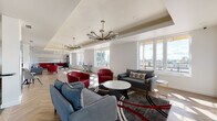 Located atop a Philadelphia building in the 19123 zip code, this common area roof lounge offers a sophisticated and well-appointed gathering space designed for comfort and socializing. The lounge is organized into distinct zones across an elongated open floor plan, all unified by warm herringbone-patterned hardwood flooring that adds a refined architectural detail throughout.

The main seating area is furnished with bold red leather club chairs, gray loveseats, and a circular coffee table arranged atop a layered area rug, creating an inviting focal point. Tray ceilings with recessed lighting are accented by a series of statement chandeliers—including a striking sputnik-style fixture and globe pendant lights with brass hardware—lending the space a contemporary yet stylish atmosphere.

Expansive windows line the exterior wall, flooding the interior with natural light and framing panoramic views of the Philadelphia skyline and surrounding rooftops. A built-in linear fireplace adds warmth to one section of the lounge.

A dedicated kitchenette area features bold red cabinetry, white subway tile backsplash, open shelving, a sink, and graphic black-and-white hexagonal floor tile, offering practical amenities for gatherings. The overall ambiance balances modern design with comfortable, communal functionality.