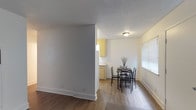 This single-story space offers a straightforward, functional layout with a clear connection between the dining area and kitchen. The dining area features wood-style laminate flooring in a warm, light tone, complemented by clean white baseboards and neutral gray and white walls that create a bright, understated atmosphere. A textured popcorn ceiling with a flush-mount light fixture provides overhead illumination, while a window fitted with horizontal blinds along the side wall allows natural light to filter into the room. A compact dining set with a dark-framed table and matching chairs anchors the dining space, making it well-suited for everyday meals. A partial wall divides the dining area from an adjacent kitchen, which features yellow-toned cabinetry and a countertop visible from the dining space. The kitchen's cabinetry provides ample storage, contributing to the overall practicality of the layout. The flooring transitions subtly between the two areas, helping to visually define each zone while maintaining a cohesive flow throughout the space. The overall ambiance is clean, modest, and functional.