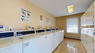This shared laundry facility is designed with functionality and convenience in mind, offering a clean and organized environment for everyday laundry needs. The single-floor space features light-toned tile flooring throughout, providing a bright and easy-to-maintain surface that contributes to the room's tidy appearance.

A prominent row of Speed Queen commercial top-load washers lines one wall, each equipped with digital card-payment terminals, making cashless transactions straightforward and efficient. Instructional signage posted above the machines guides users through proper washing and drying procedures. Adjacent to the washers, a bank of stacked commercial dryers provides ample drying capacity, maximizing the use of available space.

Natural light filters into the room through a window fitted with horizontal blinds, complemented by overhead fluorescent lighting that ensures consistent illumination throughout the day. A sturdy wooden folding table with a stone-style surface is conveniently positioned near the entrance, offering a dedicated area for sorting and folding laundry.

The facility is well-maintained and safety-conscious, with a fire extinguisher, an exit sign, and a security camera all clearly visible. A glass-panel entry door opens to the exterior, providing views of the surrounding area and enhancing ventilation. A waste bin is also available for added convenience. Service contact information is posted near the entrance for easy reference.