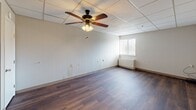 Located at 5300 Hawthorne Rd in Roanoke, Virginia, this studio unit offers an efficient, single-floor layout that includes a living room, kitchen, bathroom, and closet. The living area is finished with warm-toned hardwood-style flooring that runs throughout, complemented by a drop-panel ceiling and a ceiling fan with integrated lighting — providing both comfort and functionality. Neutral-toned wall paneling gives the space a clean, understated look, while a window along one wall brings in natural light and adds a sense of openness. The kitchen area is equipped with a full-size refrigerator and countertop workspace, keeping essentials within easy reach. A dedicated closet with a wooden door provides convenient storage. The bathroom features a full soaking tub and shower combination with crisp white surround walls, built-in shelving niches, and stainless steel grab bars — designed with accessibility and safety in mind. A ceiling vent ensures proper ventilation throughout. The overall layout is thoughtfully organized to maximize the use of the available space, making it a practical and functional unit suitable for everyday living.
