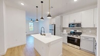 Located on Constitution Ave in Colorado Springs, CO, this well-appointed one-bedroom, one-bathroom residence offers a thoughtfully designed single-level layout. The open-concept kitchen and living area serves as the heart of the home, anchored by a striking white kitchen island with a built-in sink and matte black faucet, illuminated by a trio of pendant lights with brass accents. The kitchen features crisp white shaker cabinetry, a hexagonal tile backsplash, stainless steel appliances including a refrigerator, range, and microwave, and light wood-tone luxury vinyl plank flooring that flows throughout the entire space. A ceiling fan adds comfort to the living area, which benefits from natural light through a window overlooking greenery. The bedroom is a bright, serene retreat with multiple windows framing outdoor views, a ceiling fan, and continued wood-tone flooring. A private hallway connects the bedroom to the full bathroom, which is finished with gray cabinetry, a sleek LED-lit vanity mirror, matte black hardware, and tile accent walls. A dedicated closet and in-unit laundry area complete this efficiently designed residence, offering modern comfort and style throughout.