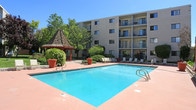 Located at 3901 Indian School Rd NE, this outdoor communal area serves as a central amenity for the residential complex. The centerpiece is a generously sized swimming pool with clear turquoise water, framed by a wide terracotta-toned pool deck that provides ample space for lounging. Multiple poolside chairs are arranged along the deck, offering plenty of seating for residents to relax and enjoy the sunny New Mexico climate.

Surrounding the pool, lush landscaping adds vibrant greenery to the space, including mature trees, flowering shrubs, and ornamental potted plants that lend a welcoming, well-maintained atmosphere. A distinctive red-painted wooden gazebo with a rustic shingled roof serves as a shaded gathering spot nestled among the trees, connected to the pool area by a concrete walkway.

The pool area is enclosed within the courtyard of a multi-story residential building, whose balconied facades are visible on multiple sides, providing a sense of privacy and community. Posted pool rules indicate structured hours of operation and access is restricted to current residents with a valid pool pass, ensuring a secure and orderly environment. Safety equipment, including a life ring, is stationed on-site. The overall ambiance is bright, open, and resort-like, making it an inviting outdoor retreat.