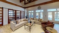 The Silver Oaks clubhouse is a thoughtfully designed single-floor community space that combines rustic architectural charm with modern amenities. The centerpiece of the space is a dramatic great room featuring soaring vaulted ceilings adorned with exposed wood beam trusses and track lighting, creating a warm yet airy atmosphere. A floor-to-ceiling stone fireplace anchors one wall, topped with a large mounted television, while rich hardwood-style flooring runs throughout the space. The lounge area is furnished with plush neutral sofas, leather accent chairs, and a glass-top coffee table, offering a comfortable setting for socializing or relaxing. Sliding barn-style doors with wood-framed glass panels add a refined rustic touch and allow natural light to flow throughout. A dedicated kitchenette area, branded with the Silver Oaks logo, features dark cabinetry with white countertops, a dual-basin sink, open shelving, a stainless steel refrigerator, and subway tile backsplash — well-equipped for hosting and everyday resident use. A wall of large glass doors and transom windows lines one side of the space, flooding the interior with natural light and providing direct views and access to the outdoor pool and patio area, seamlessly connecting indoor and outdoor living.