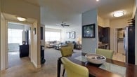 This single-floor residence features two bedrooms and two bathrooms within a generously proportioned floor plan designed for comfortable everyday living. The open-concept layout connects the living, dining, and kitchen areas seamlessly, creating a cohesive and functional space ideal for both relaxing and entertaining.

The living room is carpeted and anchored by a ceiling fan, providing comfort and airflow. Soft, neutral wall tones are accented by a cool blue-gray feature wall, adding a modern touch to the interior. A wall-mounted flat-screen television is integrated into the accent wall, contributing to a clean, streamlined aesthetic. The space receives natural light from windows along the far wall, brightening the room throughout the day.

The dining area transitions naturally from the living room, featuring a dark wood dining table paired with green upholstered chairs, providing a stylish yet approachable setting for meals. Adjacent to the dining space, the kitchen features dark cabinetry and appears well-equipped with ample storage. The textured ceiling and recessed lighting add subtle architectural character throughout the space.